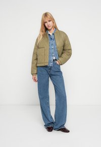 Φουσκωτό μπουφάν bomber σε ελιά πράσινο με δύο μπροστινές τσέπες, φορεμένο πάνω από ένα ανοιχτό μπλε τζιν πουκάμισο και ψηλόμεσα μπλε τζιν παντελόνια.