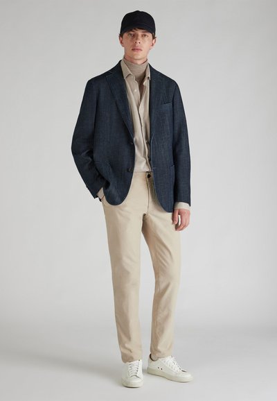 Blazer texturizado azul oscuro sobre un suéter de cuello alto beige, combinado con pantalones beige claros y zapatillas blancas. El modelo lleva una gorra oscura.