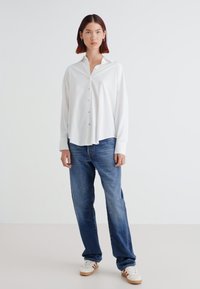 Valkoinen pitkähihaisen napillisen paidan, jossa on kaulus, on yhdistetty sinisiin löysiin farkkuihin ja valkoisiin tennareihin. Tyylikäs kangas, jossa on sileä pinta.