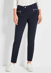 Marineblauwe slim-fit broek met twee voorzakken met ritssluiting en een gladde textuur. Gecombineerd met witte sneakers voor contrast.