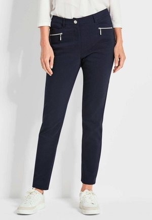 Marineblauwe slim-fit broek met twee voorzakken met ritssluiting en een gladde textuur. Gecombineerd met witte sneakers voor contrast.