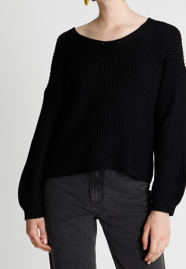 Femme portant un pull en maille côtelée noire avec de longues manches ballon et un jean noir délavé, debout devant un fond uni.