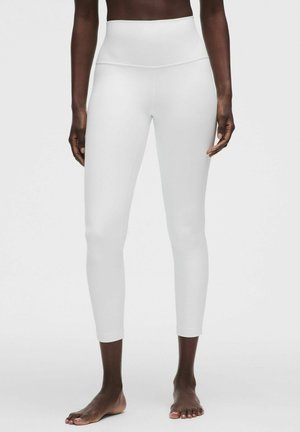 Person trägt hochtaillierte, knöchellange weiße Leggings und steht barfuß auf einem schlichten hellen Hintergrund.