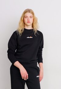 Sudadera negra con cuello redondo y logotipo en el pecho, combinada con pantalones negros. La tela parece suave y de estilo casual.