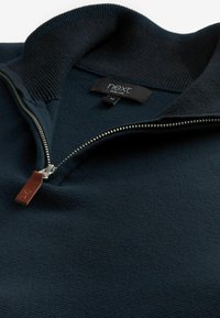 Marineblauwe zip-up trui met een gestructureerde stof, geribbelde kraag en zilveren metalen rits. Lederen trekbandje bevestigd aan de rits. Merkenlabel zichtbaar.