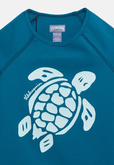 Μπλε κορυφή προστασίας από τον ήλιο με μεγάλο λευκό γραφικό χελώνας θάλασσας και το λογότυπο "Vilebrequin" εκτυπωμένο στο μπροστινό μέρος, ετικέτα μεγέθους ορατή μέσα στο γιακά.