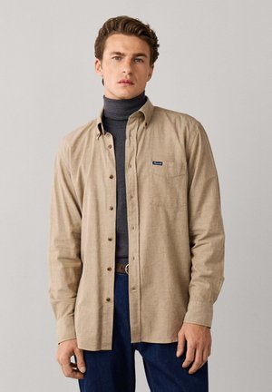 Junger Mann mit kurzen braunen Haaren trägt ein beiges Hemd über einem dunkelgrauen Rollkragenpullover und blaue Jeans, steht vor einem schlichten Hintergrund.