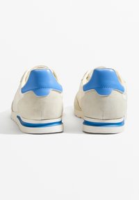 Beige színű velúr sportcipők kék bőr részletekkel, párnázott sarokkal, fehér középtalppal és texturált gumitalppal a tapadásért.