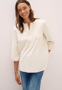 Donna sorridente con capelli castani, indossa una blusa bianca a V con maniche 3/4 e jeans blu, in piedi contro uno sfondo chiaro e semplice.