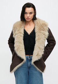 Καφέ εξωτερικό μπουφάν με γιακά από ανοιχτό μπεζ τεχνητό γούνινο ύφασμα. Υφασμα με υφή, ανοιχτό μπροστινό μέρος και άνετη εφαρμογή, σε συνδυασμό με μαύρο τοπ και μπλε τζιν.