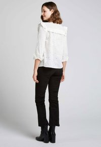 Blouse blanche à manches longues avec un design en œillets et un col volanté, associée à un pantalon noir ajusté et des bottines noires.