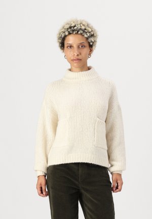 Pull en maille de couleur crème avec un col haut, deux poches avant et des bordures côtelées. Porté avec un pantalon en velours côtelé vert foncé.