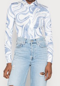 Person wearing a long-sleeve white shirt with blue swirling patterns and light blue high-waisted ripped jeans.