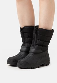 Botas de invierno negras aisladas con cremallera frontal, suelas de tacos gruesos, usadas sobre piernas desnudas, mostradas sobre un fondo blanco.