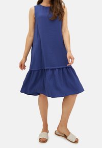 Sininen hihaton mekko, jossa on teksturoitu yläosa ja kerroksellinen röyhelöhelma, yhdistetty vaaleisiin, matalapohjaisiin slip-on-sandaaleihin.