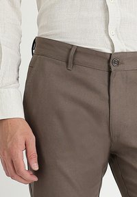 Man wearing brown trousers and a beige textured button-up shirt, with left hand relaxed by his side.