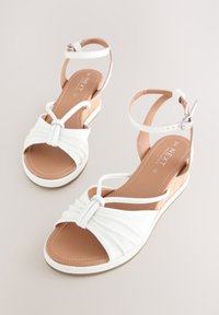 Sandales plates blanches avec un design en cuir froncé, des lanières fines et une bride de cheville à boucle. La semelle présente un discret accent en liège.