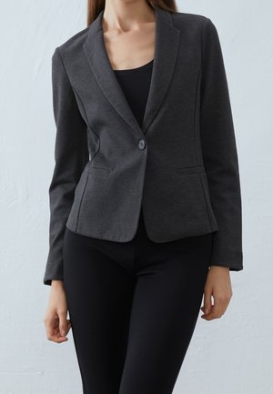 Vrouw draagt een getailleerde donkergrijze blazer met één knoop, gecombineerd met zwarte broek, staand tegen een effen lichte achtergrond.