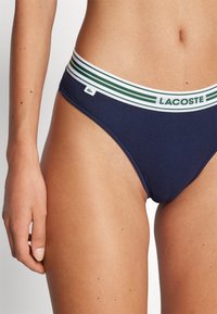 Tummansininen puuvillabikinihousu, jossa on valkoisen ja vihreän raidallinen vyötärö, jossa on "LACOSTE"-logotagi sivussa.