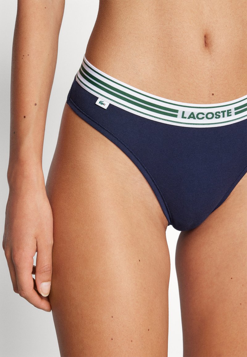 Bas de bikini en coton bleu marine avec une ceinture rayée blanche et verte portant l'étiquette logo "LACOSTE" sur le côté.