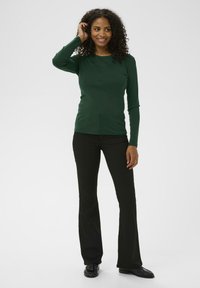 Chemise longue à manches longues vert foncé avec un col rond, assortie à un pantalon noir évasé et des chaussures noires, présentant une texture lisse.