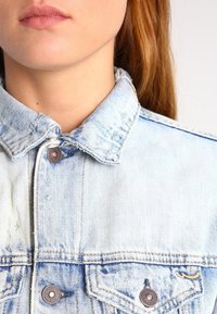 Light blue denim jacket with a pointed collar, visible metal buttons, and distressed detailing. Fabric texture is soft with faded patterns.
