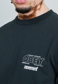 Black cotton t-shirt featuring a white graphic print with the text "OBEY PERPETUAL PERSISTENT movement" on the left chest area.