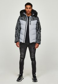 Giacca imbottita grigia con maniche nere a motivo, rifinitura in pelliccia sintetica sul cappuccio e dettaglio del logo. Indossata con jeans neri strappati e sneakers.