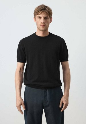 Hombre con pelo corto y rubio, que lleva una camiseta negra de manga corta y pantalones azul oscuro, de pie frente a un fondo liso y claro.