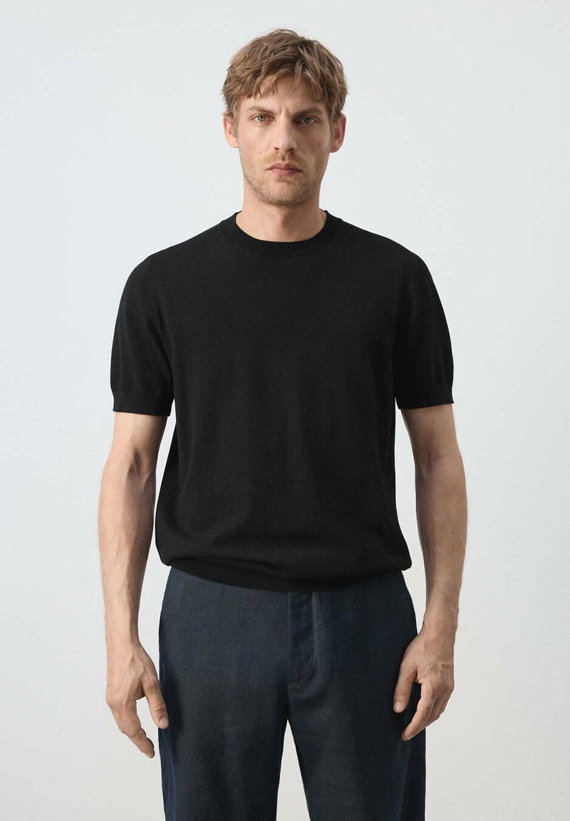 Homme aux cheveux blonds courts portant une chemise noire à manches courtes et un pantalon bleu foncé, se tenant devant un fond clair uni.