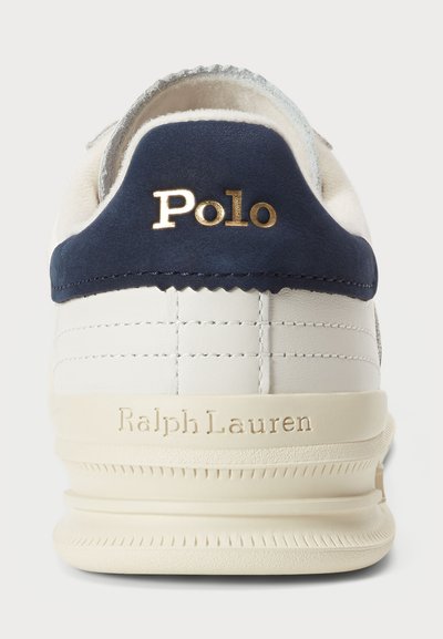 White sneaker with navy suede heel, gold "Polo" logo, textured leather, and light rubber sole featuring horizontal grooves and "Ralph Lauren" embossed.