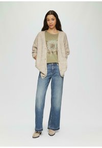 Gilet beige, t-shirt graphique vert avec un motif en éclosion solaire, et jeans évasés bleu clair. Texture douce et fermeture à boutons sur le gilet.