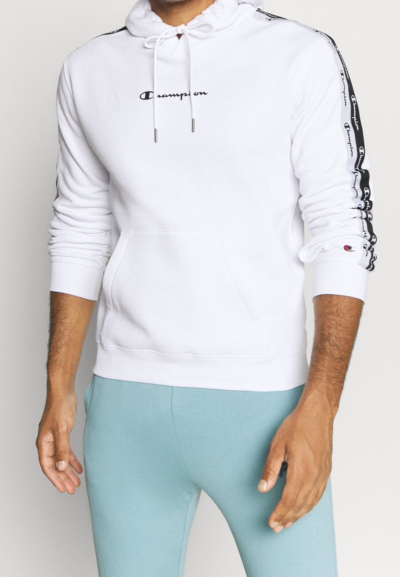 Sweat à capuche blanc avec poche avant, capuche à cordon de serrage et logo noir. Bande de logo noire et blanche courant le long des deux manches. Tissu doux.