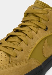 Baskets en daim jaune moutarde avec lacets assortis, perforations, semelle en caoutchouc, et un panneau latéral texturé arborant un logo.