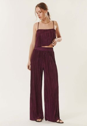 Femme portant un crop top sans manches plissé violet foncé avec un pantalon large assorti et des sandales noires, debout devant un fond uni.