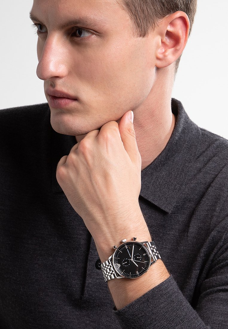 Chronographuhr mit schwarzem Zifferblatt, silbernem Edelstahlarmband und subtilen Details auf dem Zifferblatt. Tragen an einem männlichen Handgelenk, vor einem grauen Hintergrund.