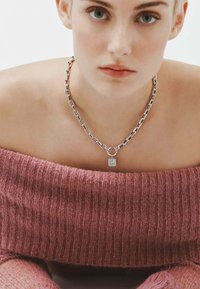 Collar de cadena plateada gruesa con un colgante rectangular, llevado sobre un suéter de punto rosa con escote off-the-shoulder y textura suave.