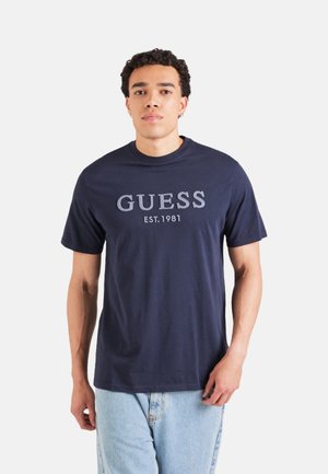 Camiseta de algodón azul marino con mangas cortas, con "GUESS EST. 1981" en texto gris claro en el delantero. Cuello redondo clásico.