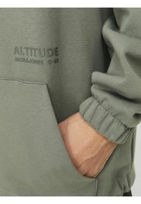 Pulover verde măsliniu cu manșete elastice și buzunar tip kanguru. Prezintă text ridicat „ALTITUDE JACK & JONES” pe față. Textură moale a țesăturii.
