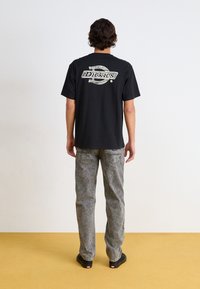 Camiseta negra de manga corta con un gran estampado gráfico blanco en la parte posterior. Se lleva con jeans grises deslavados y zapatos negros sin cordones.