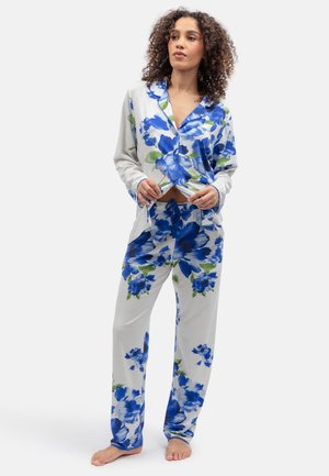 Vrouw staat blootsvoets, draagt een witte pyjama met grote blauwe en groene bloemenpatronen, een hemd met knopen en een broek met aantrekkoord tegen een witte achtergrond.