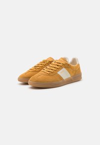 Baskets en daim jaune avec des accents blancs, bout arrondi et semelle en caoutchouc. Présente des lacets et une marque sur la languette et le côté.