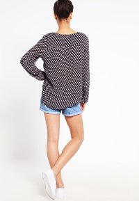 Schwarz-weiß gemusterte Bluse mit langen Ärmeln, lockere Passform, getragen mit hellblauen Jeansshorts und weißen Sneakers, von hinten betrachtet.