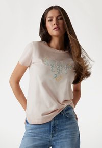 Camiseta de algodón de color rosa claro con un logo bordado "GUESS" en azul y amarillo con acentos florales. Mangas cortas y ajuste relajado.