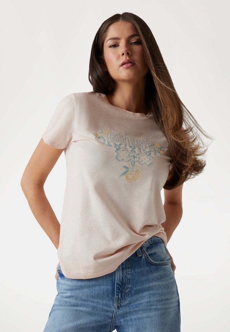 Camiseta de algodón de color rosa claro con un logo bordado "GUESS" en azul y amarillo con acentos florales. Mangas cortas y ajuste relajado.