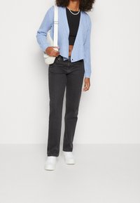 Femme portant un cardigan bleu clair, un crop top noir, un jean foncé, des baskets blanches, un collier de perles et portant un sac à bandoulière blanc.