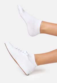 Hvide lærreds sneakers med gummisål, der har grå øjer og snørebånd, iført en hvid ankelstrømpe. Foden er placeret vandret.