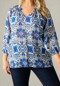 Blusa azzurra a fantasia realizzata in tessuto leggero con scollatura a V, colletto arricciato e maniche a tre quarti a sbuffo con disegni intricati.