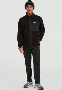 Jovem vestido com um casaco de fleece preto com fecho, calças escuras, ténis cinzas e um gorro verde a posar contra um fundo simples.