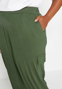 Personne portant un pantalon vert olive avec une poche latérale et une main placée dans la poche avant droite sur fond blanc.